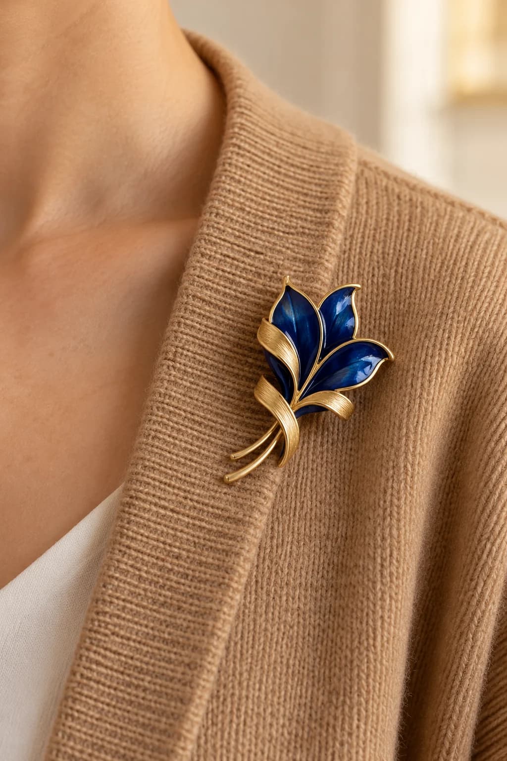 Compact blue enamel and gold brooch worn on the supported shoulder edge of a camel cardigan