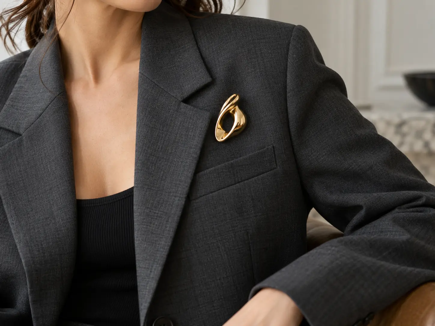 Sculptural gold brooch positioned above the breast pocket of a charcoal blazer