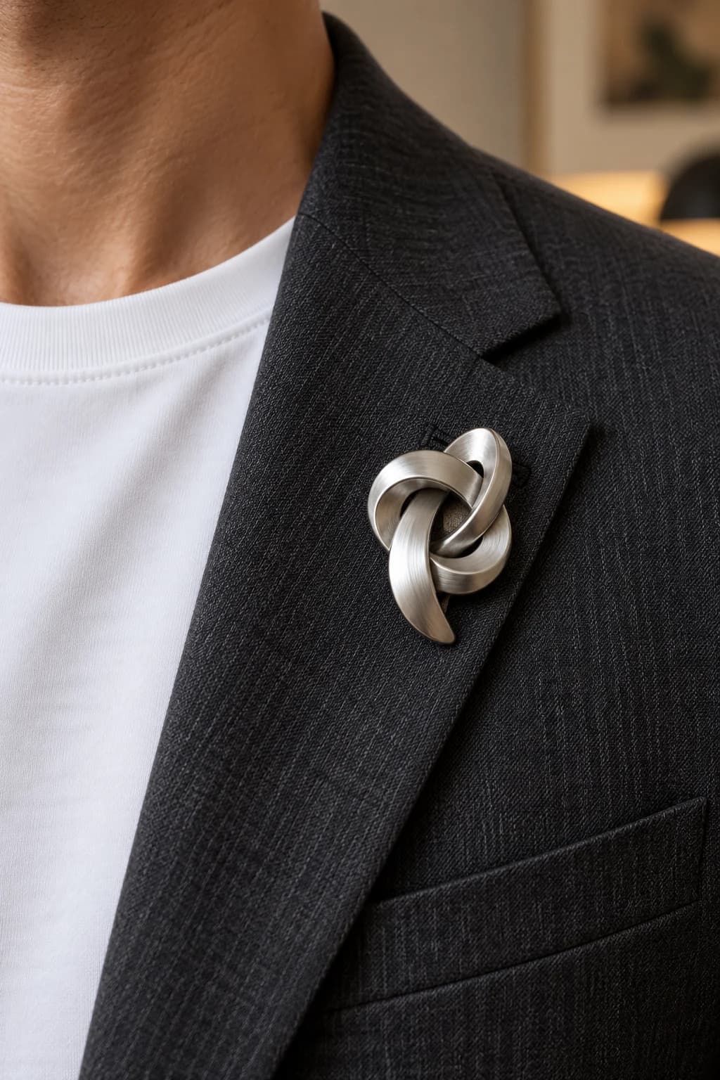 Brushed-silver knot brooch worn high on the lapel of a charcoal men's blazer over a white T-shirt