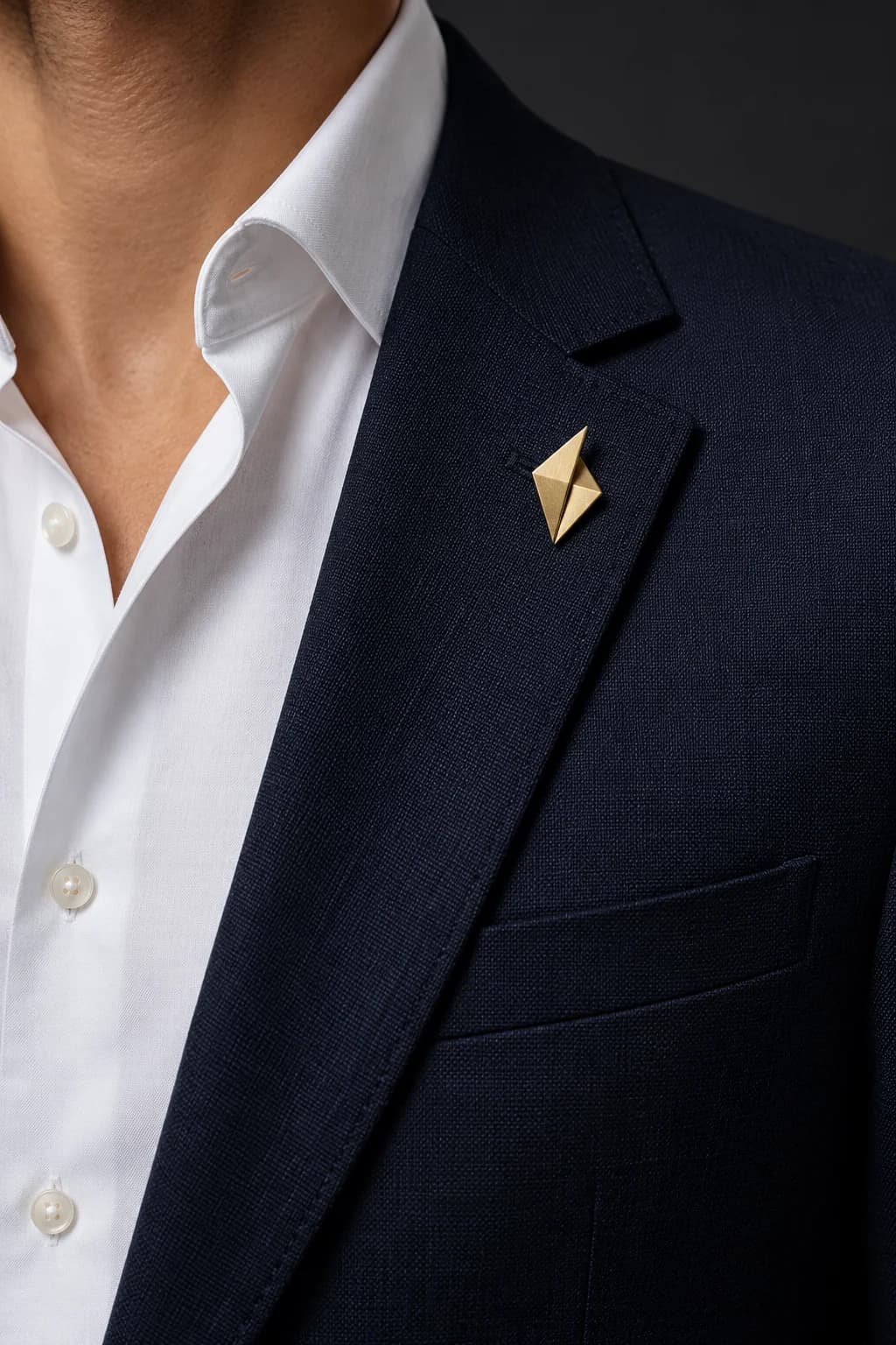 Small matte-gold geometric brooch positioned below the notch of a midnight-navy men's suit lapel