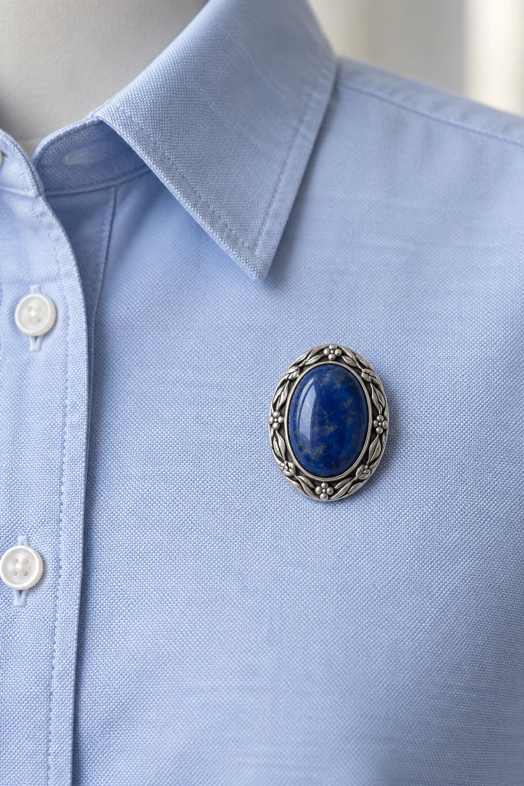 Oval silver brooch with a deep blue stone on the upper chest of a pale blue Oxford shirt