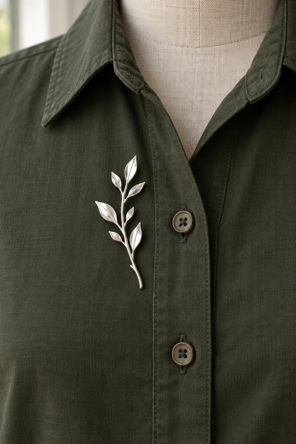 Slim silver branch brooch beside the upper button placket of a dark olive cotton shirt