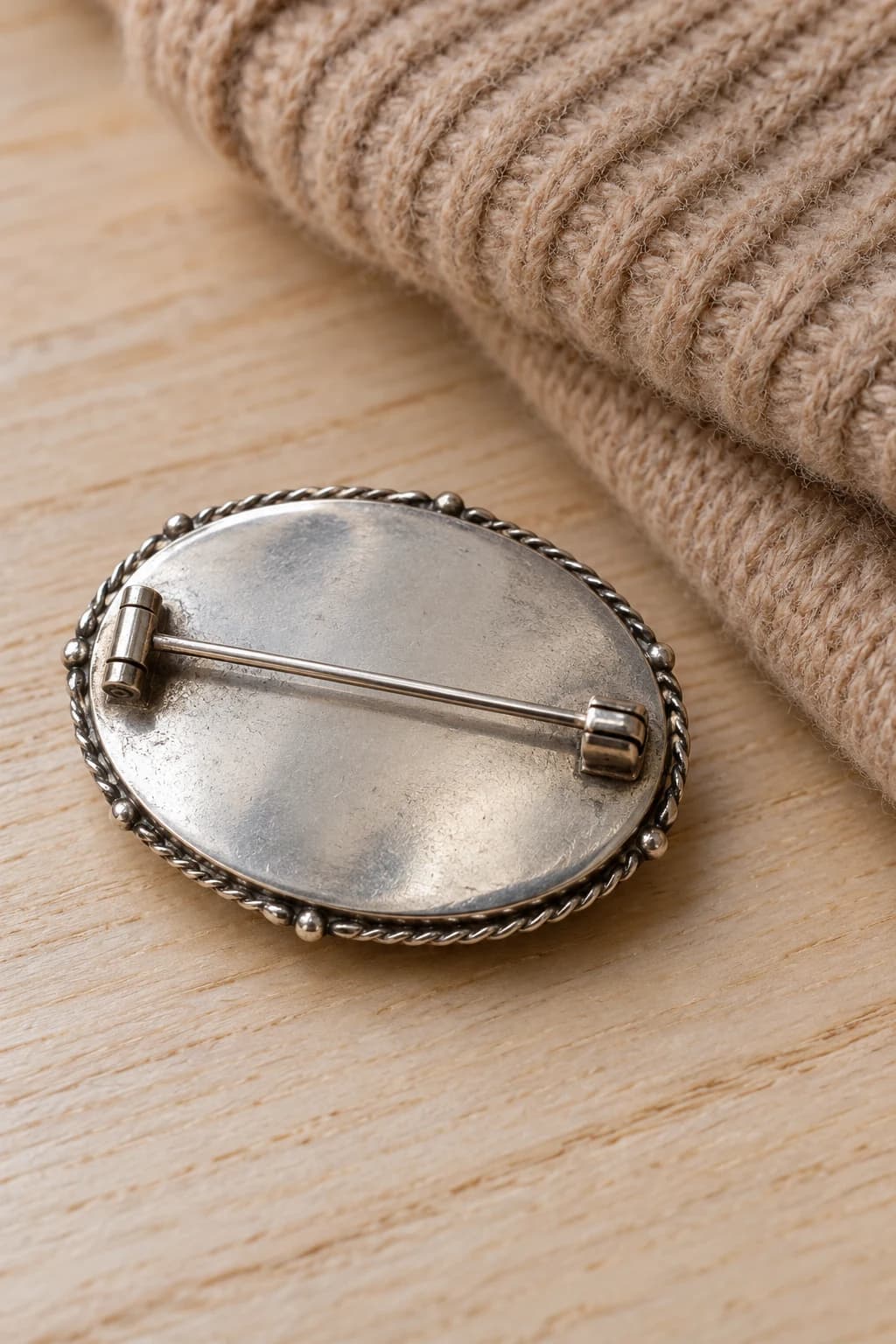 Back of a silver oval brooch showing its closed pin and safety catch beside folded camel knitwear